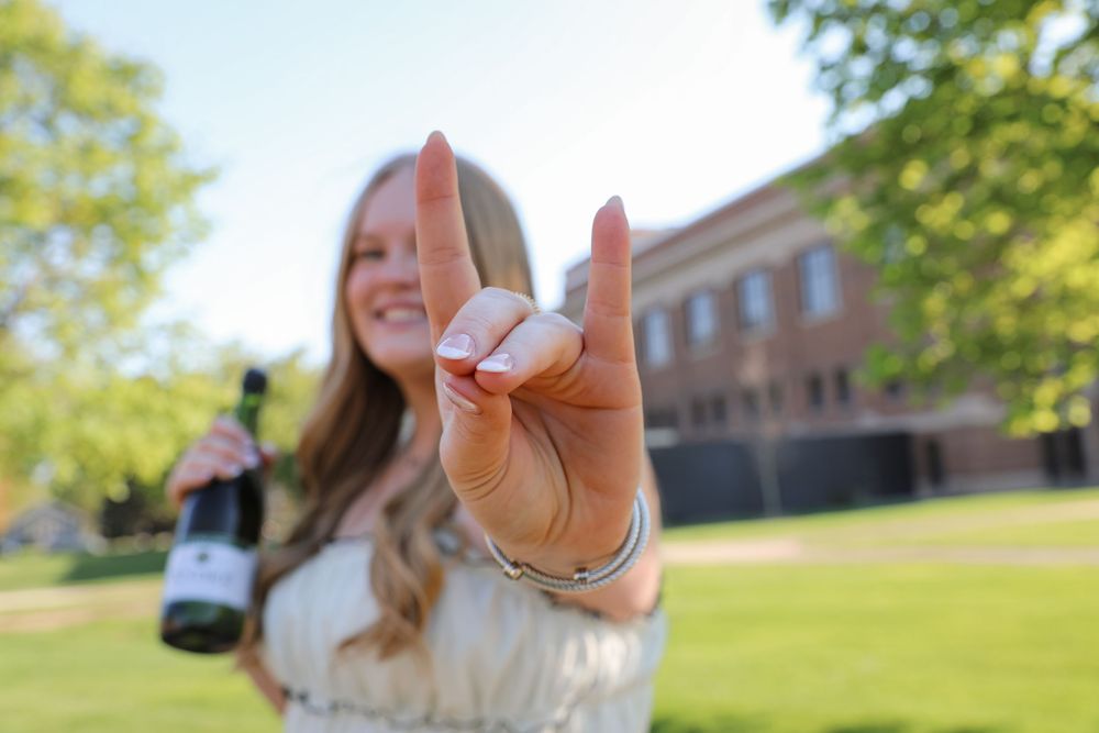 Young woman holding wine bottle making rock on hand gesture outdoors on sunny college campus.