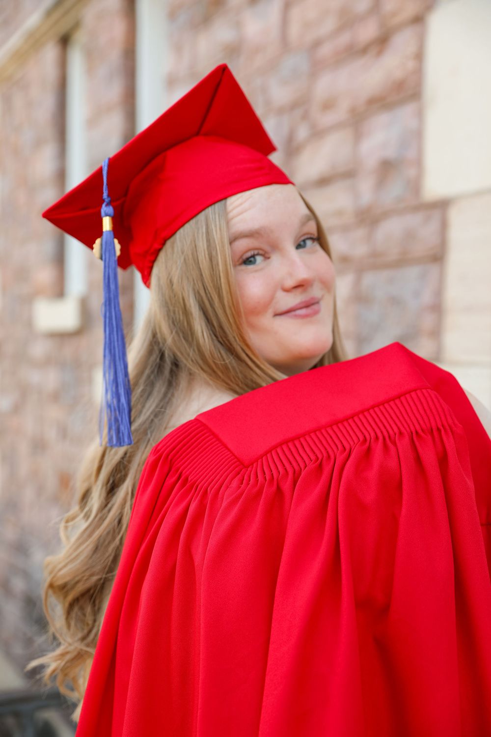 Young woman in red graduation cap and gown smiling over her shoulder outdoors near a brick building.