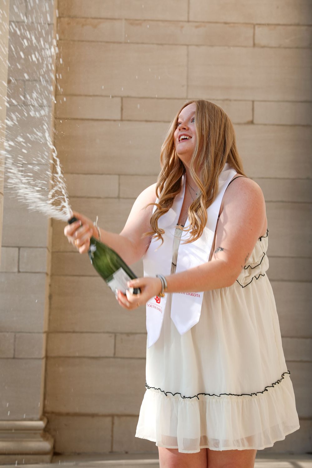 Young woman in white dress joyfully popping champagne bottle outdoors during graduation celebration.