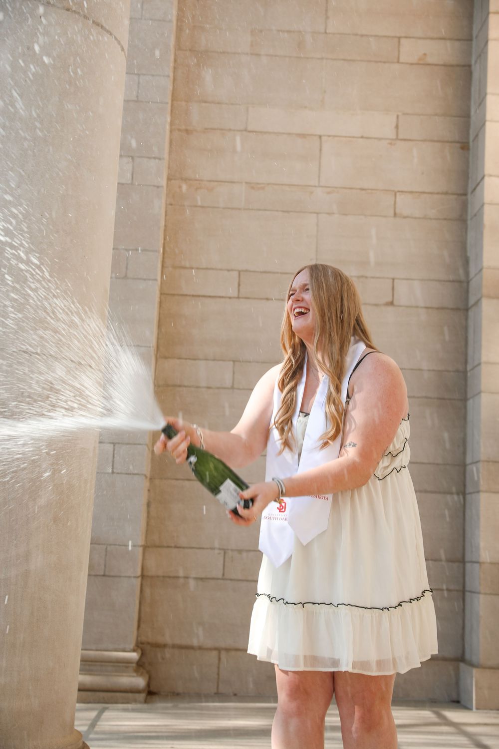 Smiling blonde woman in white dress popping champagne bottle outdoors near stone columns.