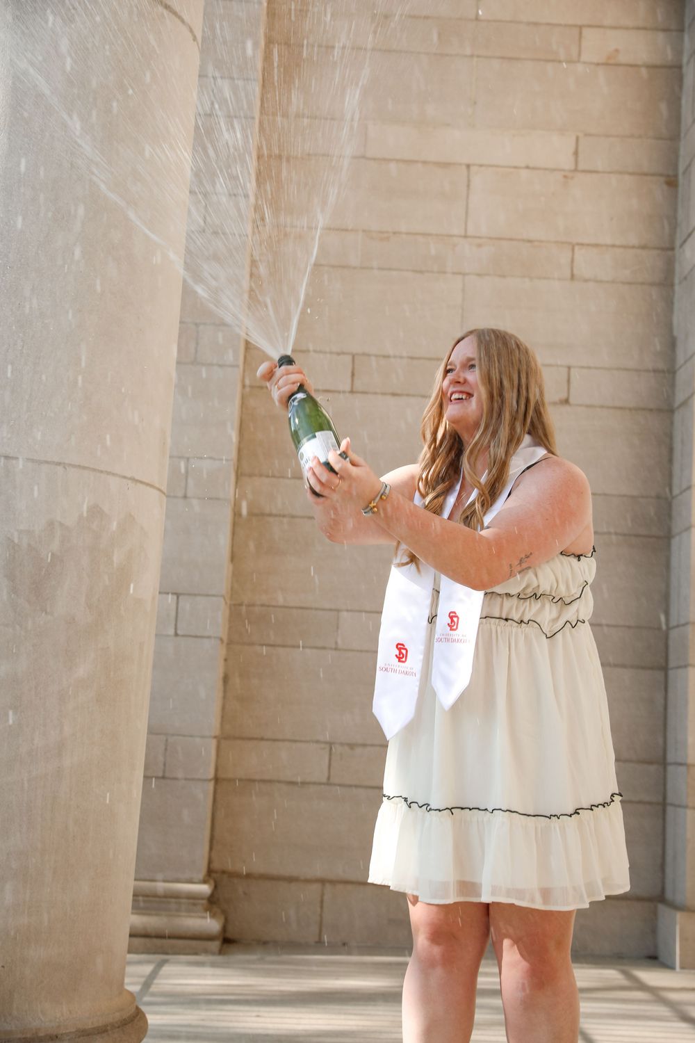 Graduate in white dress and sash joyfully sprays champagne bottle outdoors near stone columns.