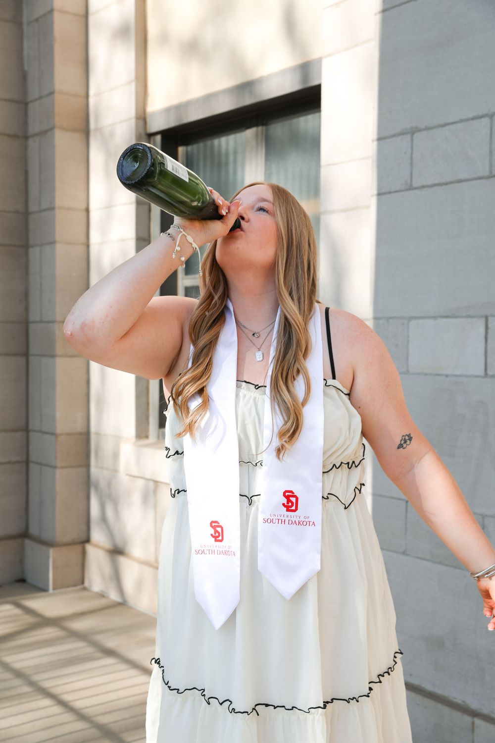 University of South Dakota graduate celebrates in white dress and SD stole, drinking champagne bottle outdoors.