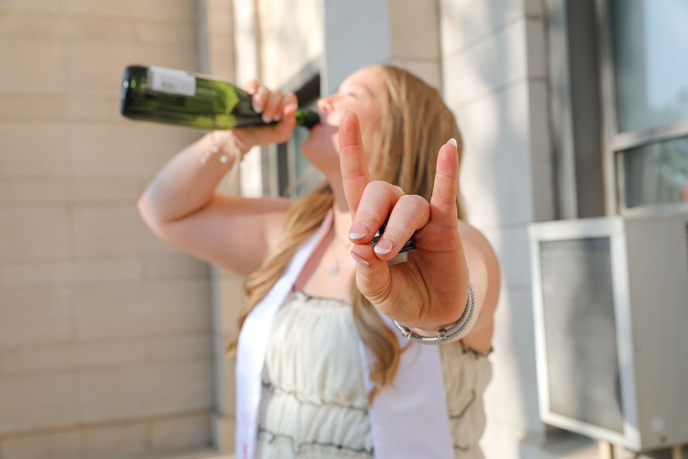 Woman drinking champagne from bottle while wearing handcuffs and making rock sign gesture outdoors.