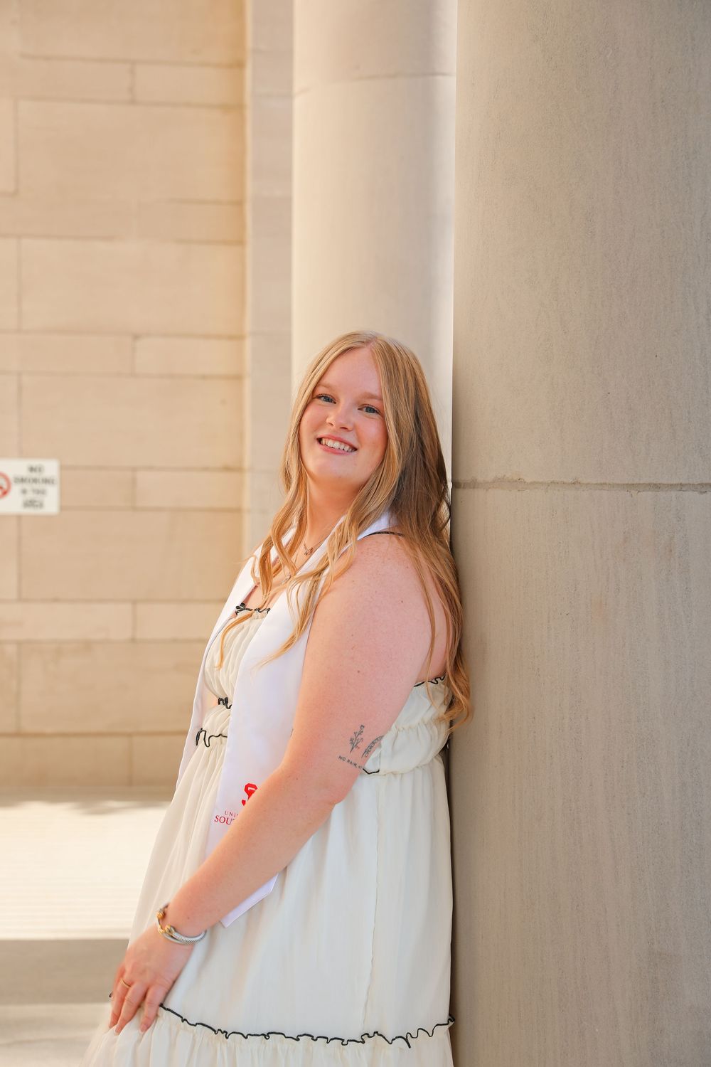 Young blonde woman in white dress leaning against stone column, smiling outdoors in warm sunlight.