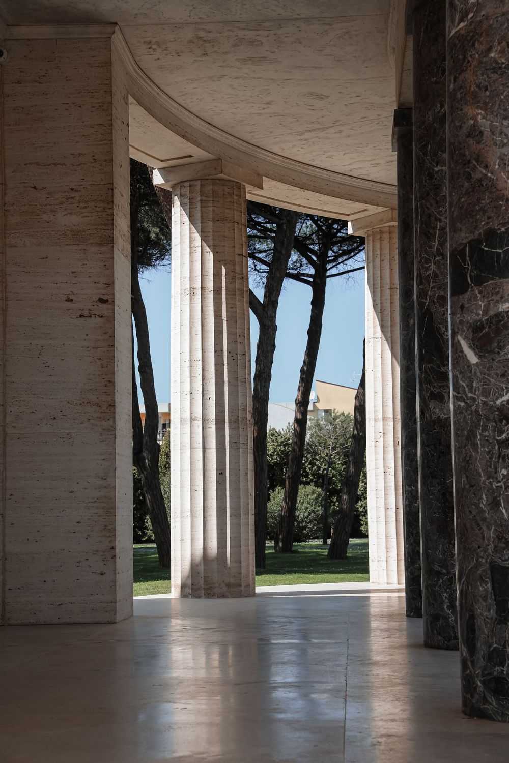 Elegant marble colonnade with fluted columns framing a sunlit garden with tall pine trees and blue sky.
