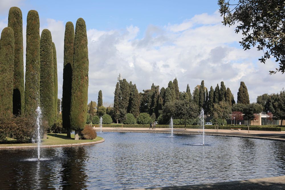Elegant garden fountain pool with tall cypress trees and water jets under a partly cloudy sky.