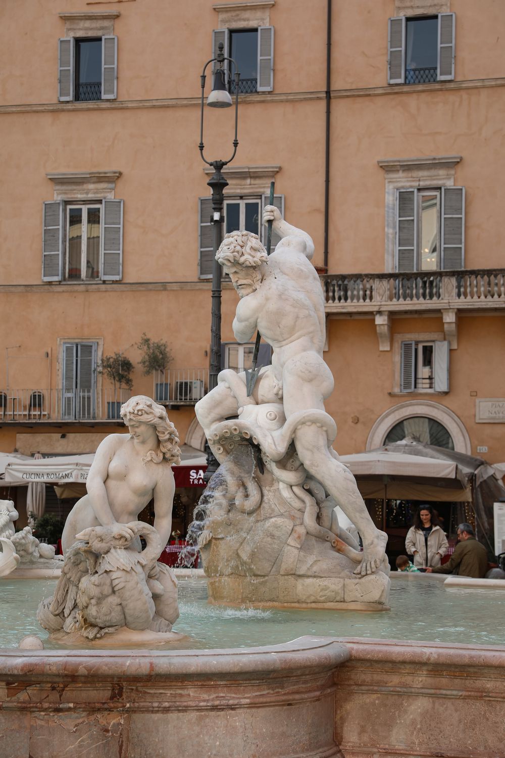 Marble fountain sculptures at Piazza Navona in Rome, Italy, with historic orange building facade in background.