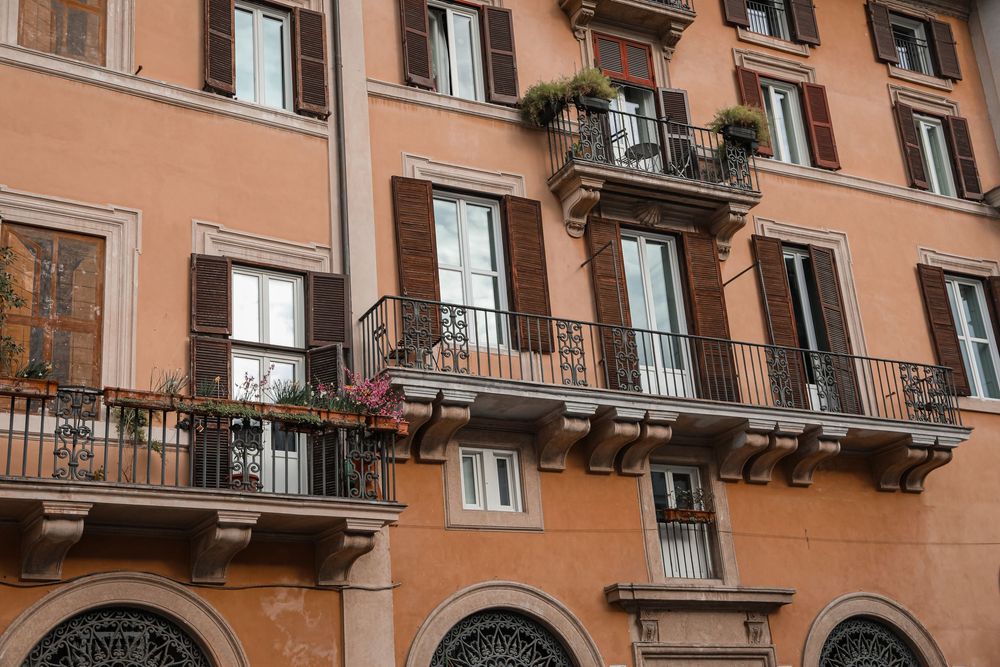 Ornate Italian building facade with wrought iron balconies, flower boxes, and brown shutters on terracotta walls.
