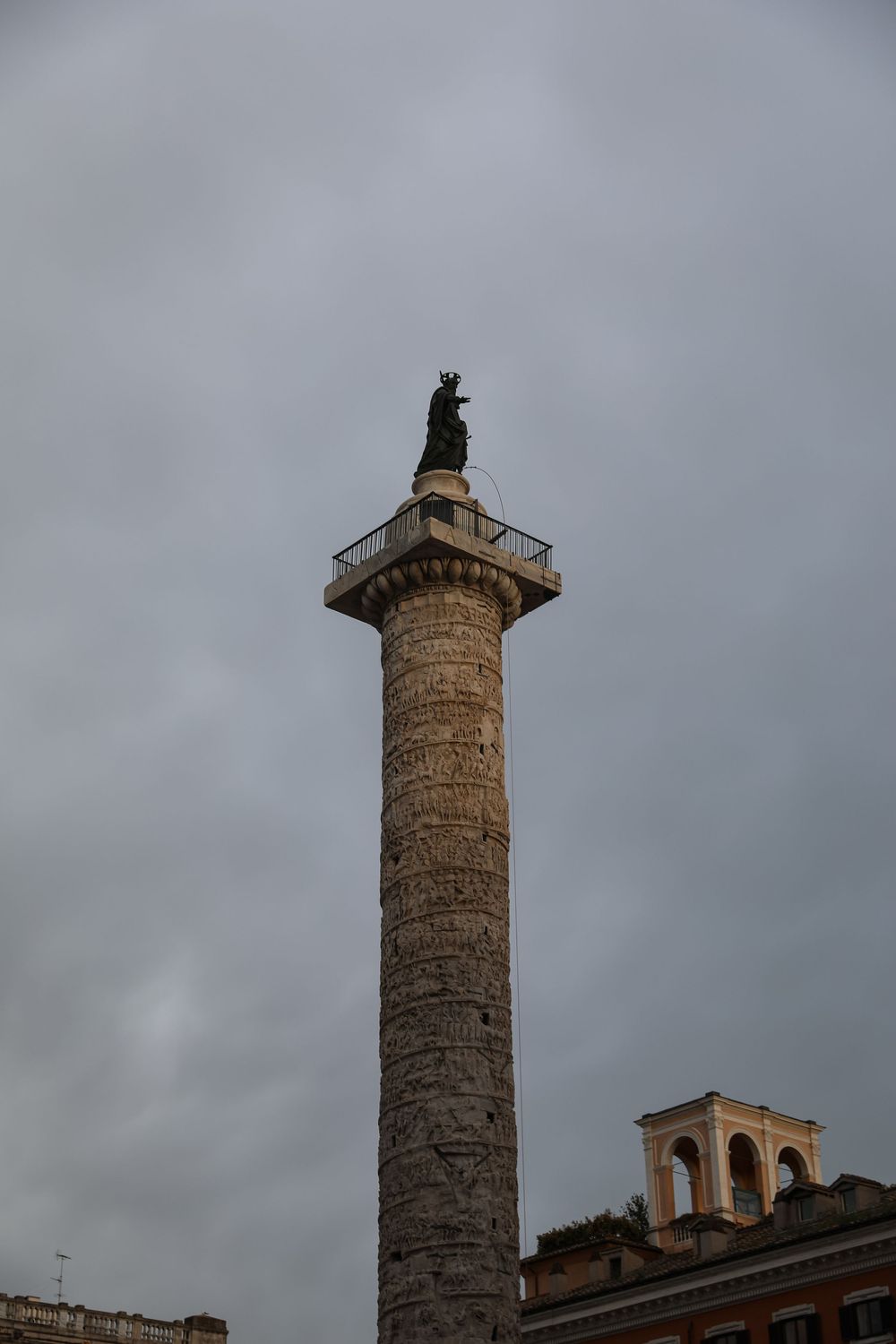 Trajan's Column in Rome, Italy, topped with a bronze statue against a dramatic cloudy sky.