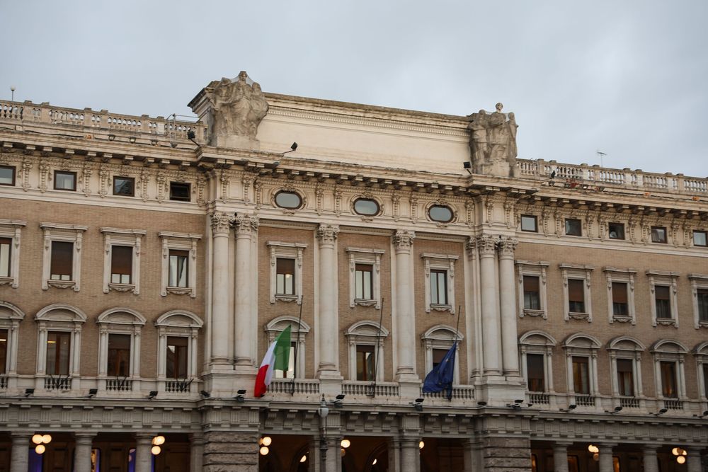 Ornate neoclassical Italian government building facade with arched colonnades, decorative sculptures, and Italian and EU flags.
