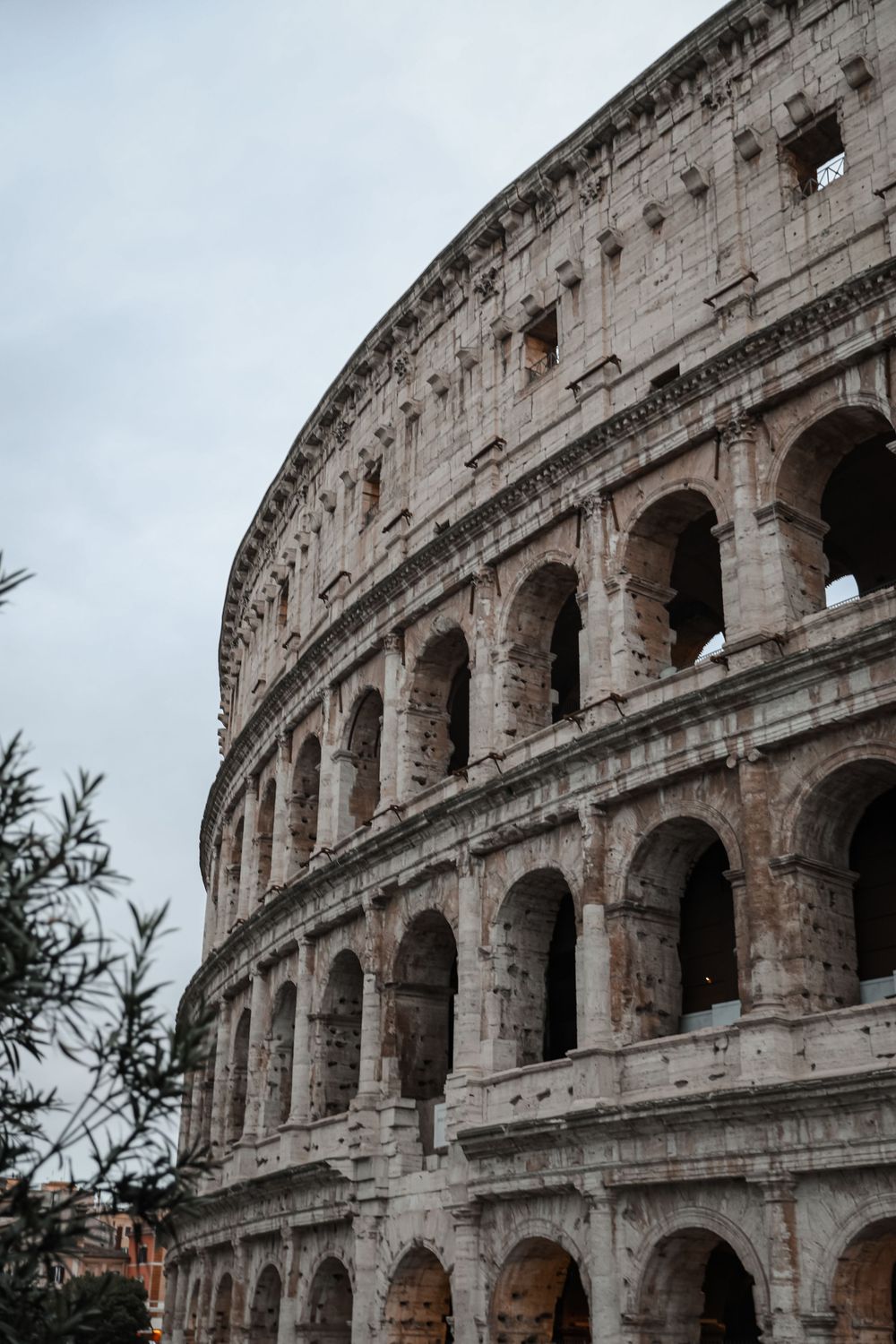 Ancient Roman Colosseum exterior with arched facades and weathered stone walls under overcast sky in Rome, Italy.