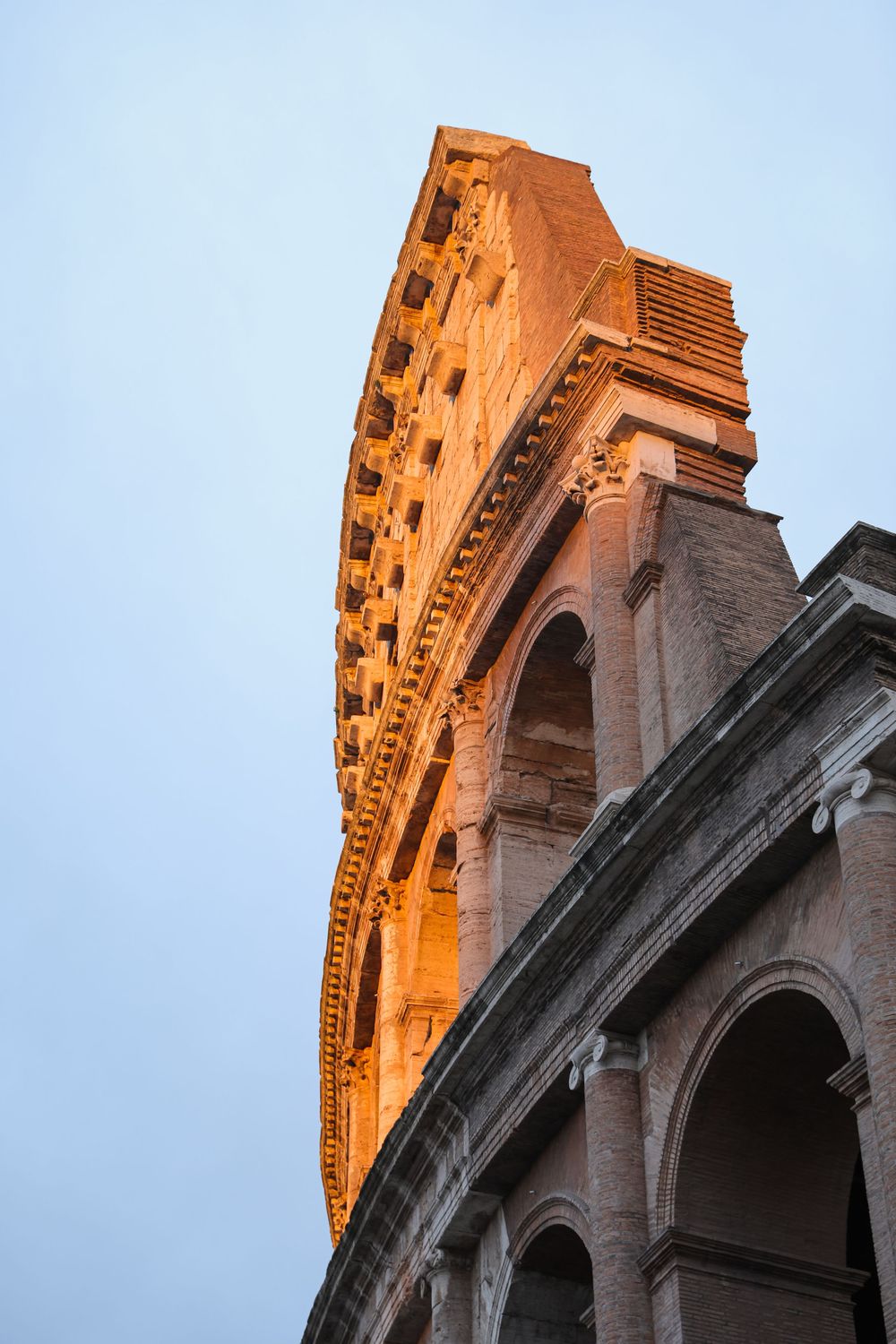 Golden-lit Colosseum exterior wall with arched openings photographed from low angle at dusk in Rome, Italy.