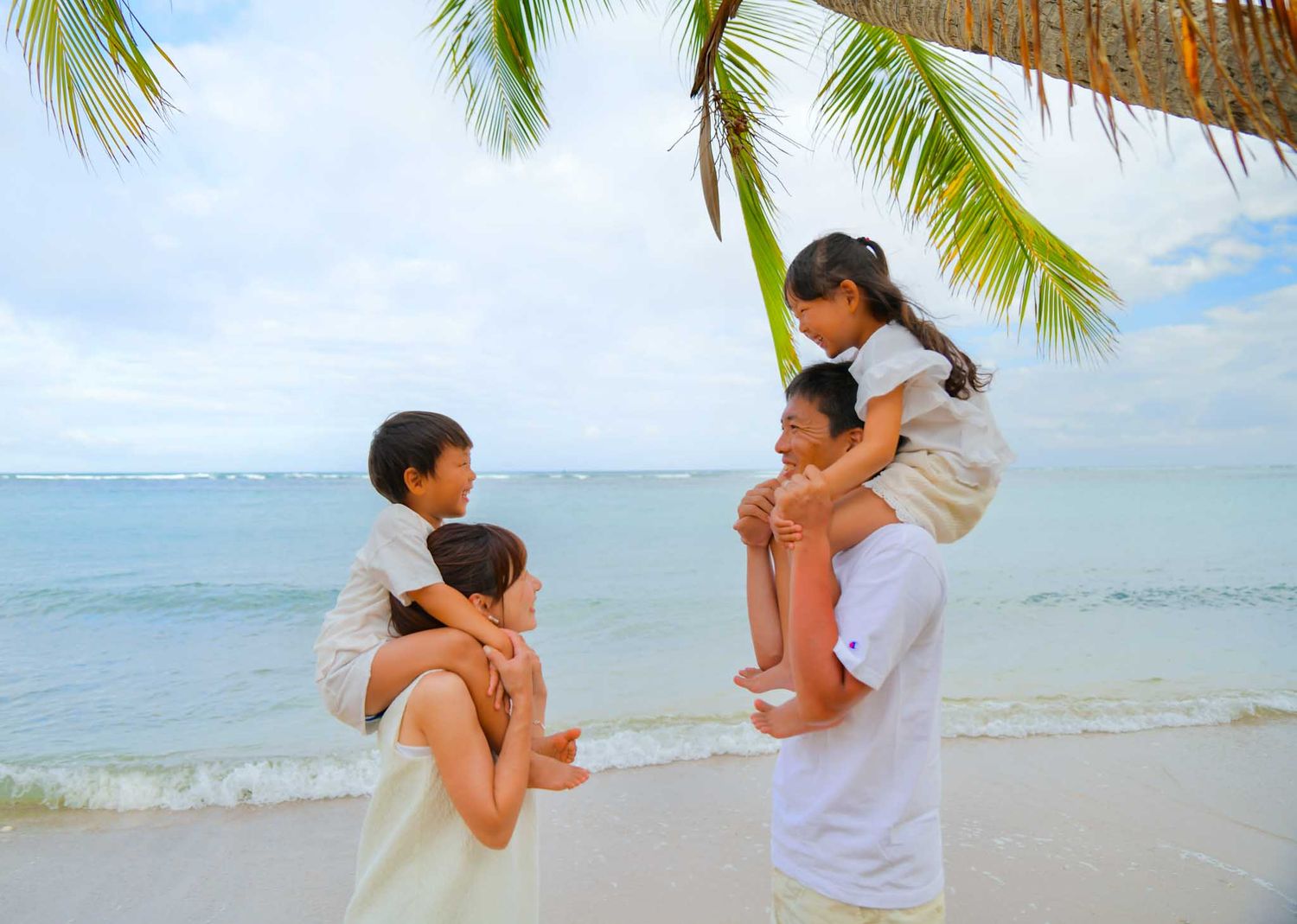 Family of four enjoying a tropical beach vacation, parents carrying children under a palm tree by turquoise ocean waters.