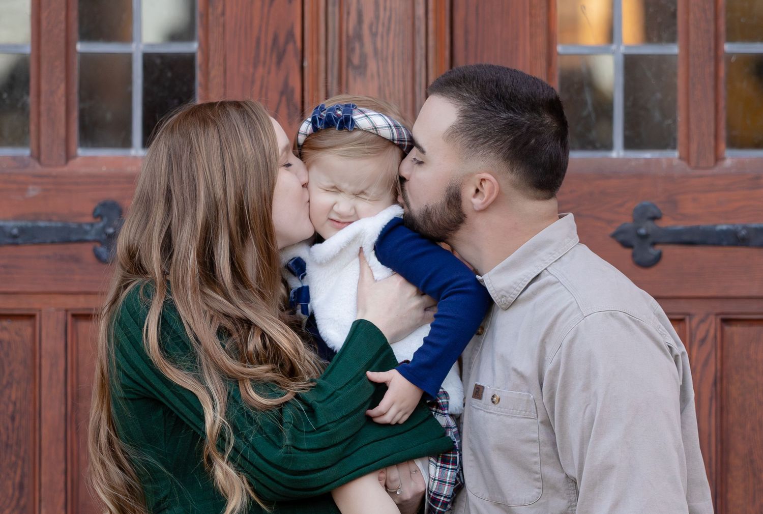 Maternity Photographer Calhoun GA, A couple kisses their baby on the cheeks in front of elegant wooden doors in a warm family portrait.