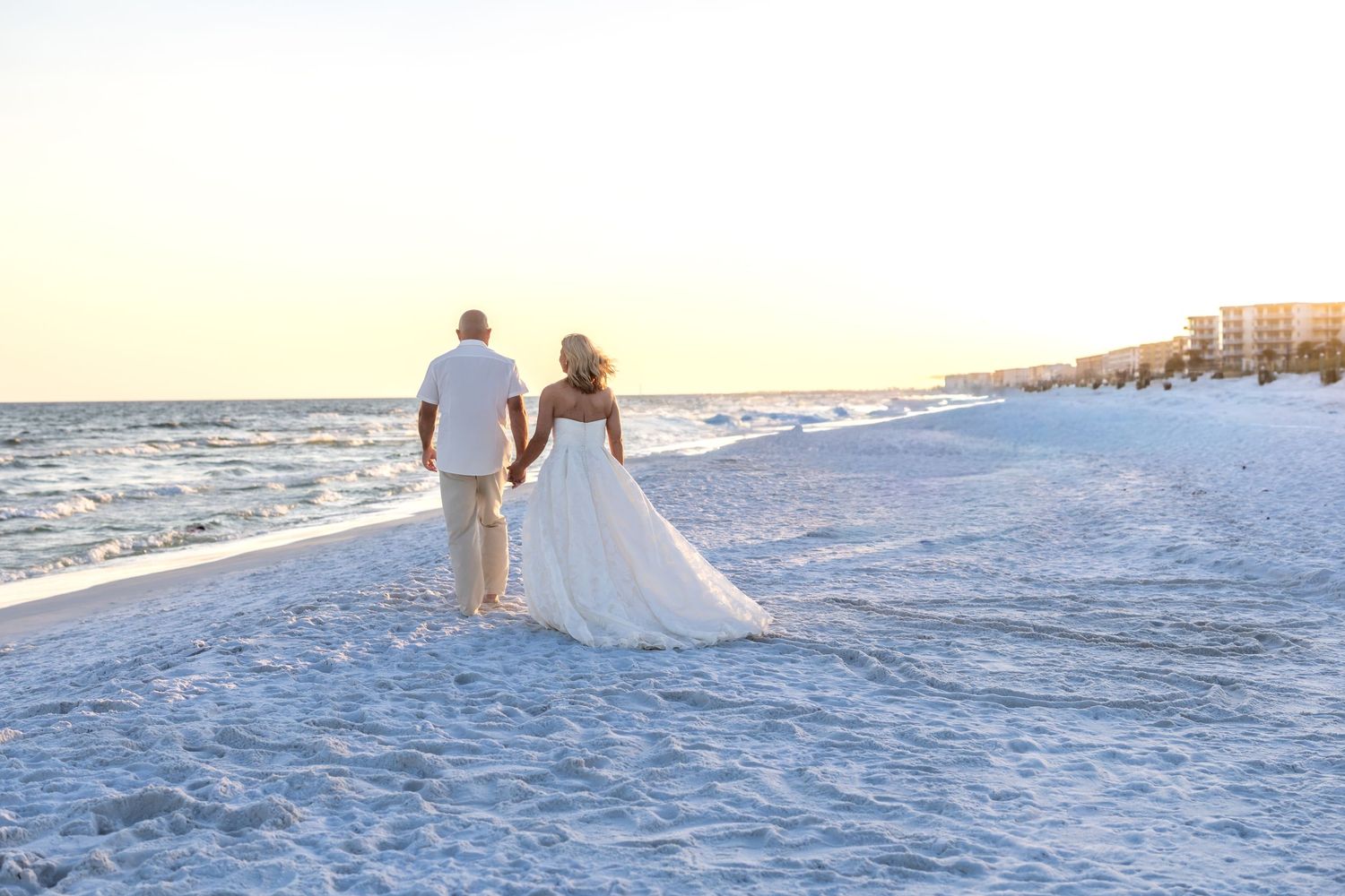 Bride and groom walking hand in hand on a white sand beach at sunset in a romantic wedding photo.