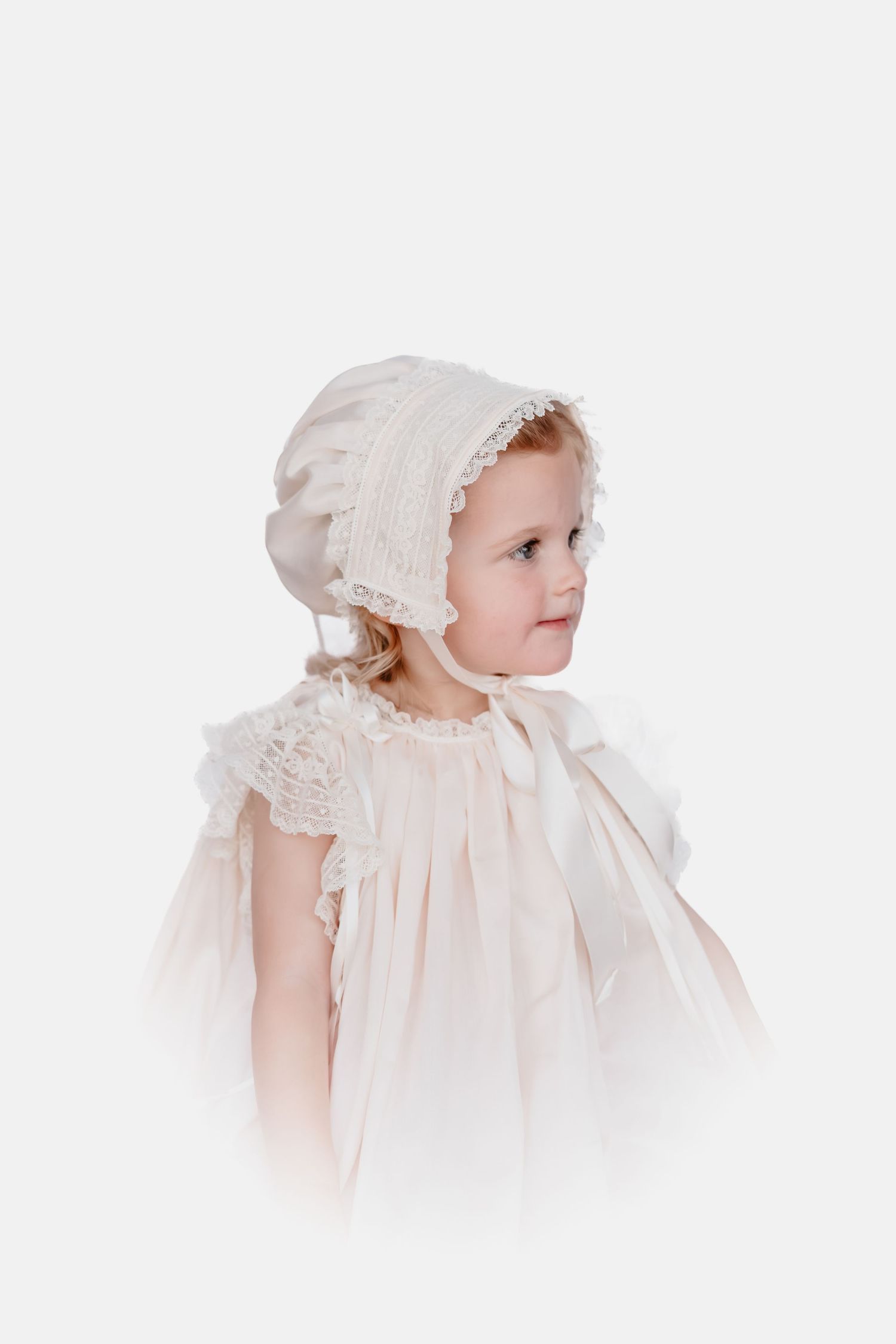 Young girl in a cream heirloom bonnet and pleated dress with lace flutter sleeves and satin ribbon trim, shown in side profile.