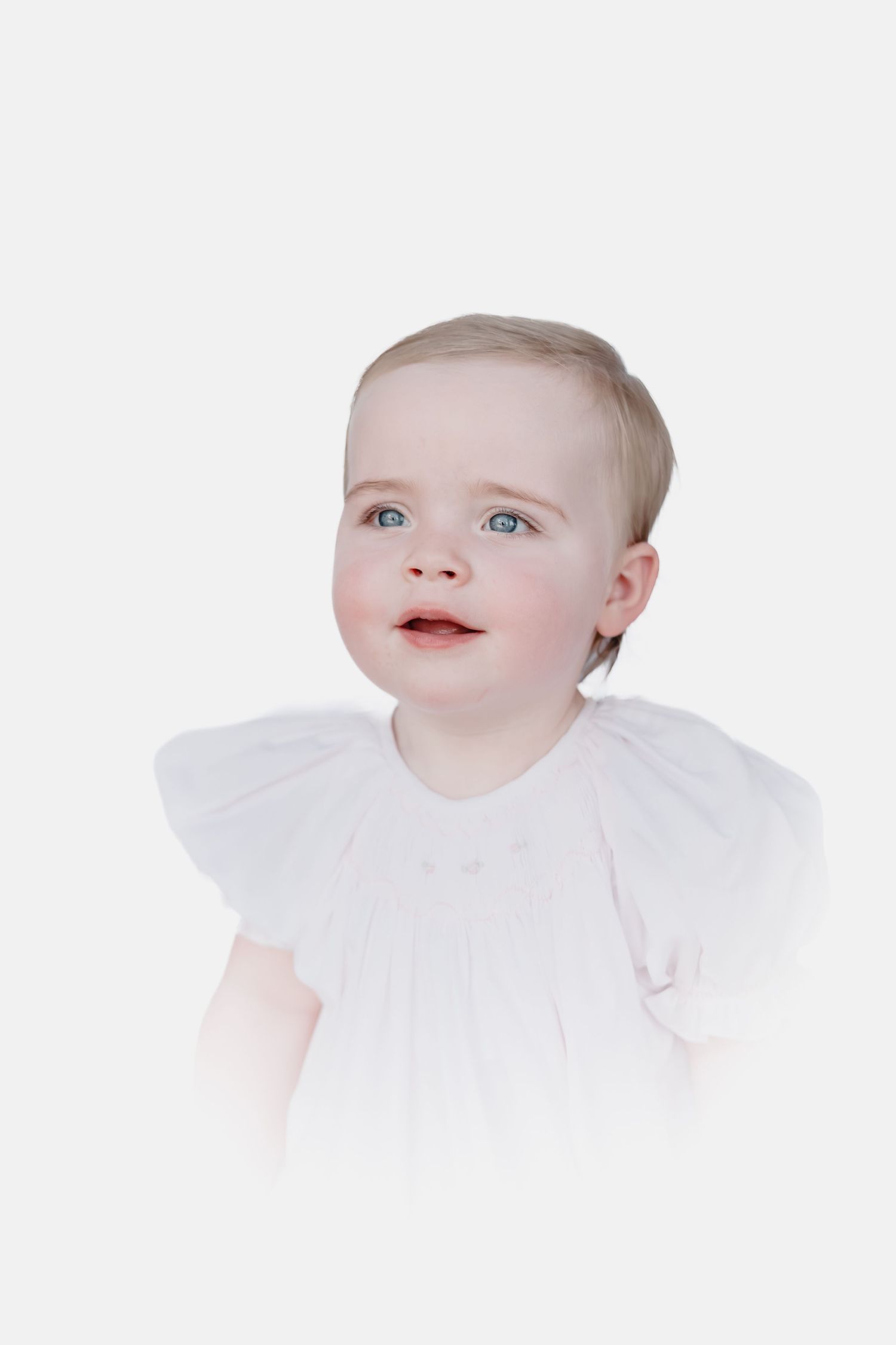 Smiling baby with blue eyes and short blonde hair wearing a white smocked dress with flutter sleeves against a soft white background.