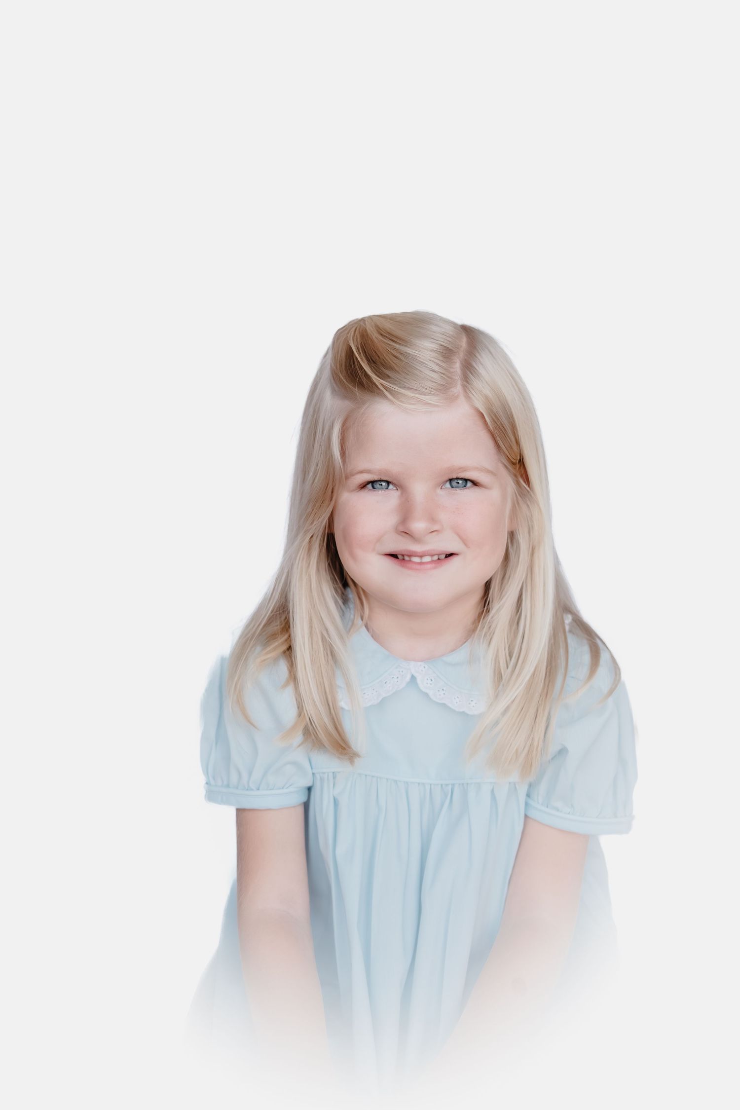 Smiling young girl with long blonde hair wearing a light blue short-sleeved dress with a collar, posed against a soft white background.