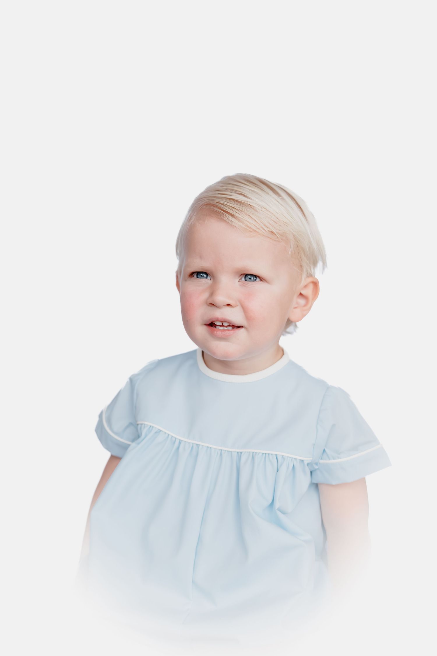 Blonde toddler with blue eyes wearing a light blue short-sleeve smocked top with white piping against a soft white background.
