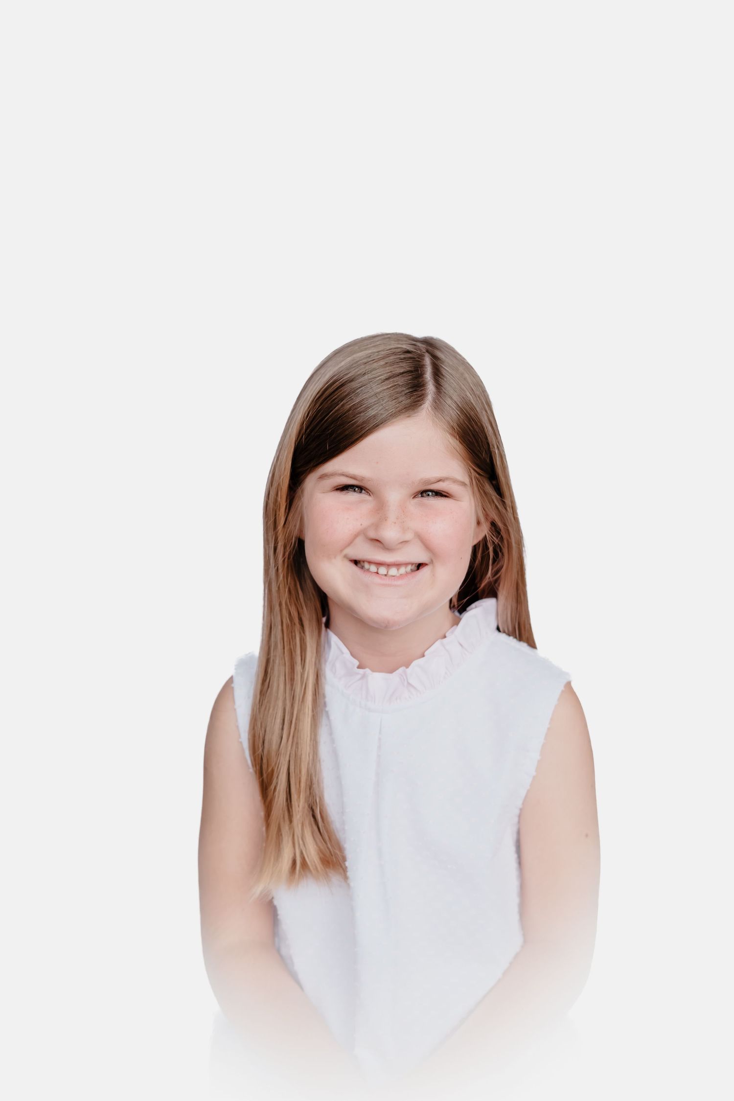 Smiling young girl with long straight brown hair wearing a white sleeveless ruffled-collar top against a soft white background.