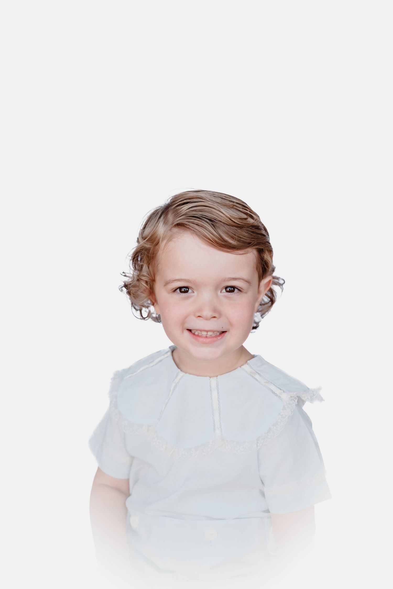 Smiling toddler with wavy light brown hair wearing a white ruffled-collar blouse in a bright studio portrait on a white background.