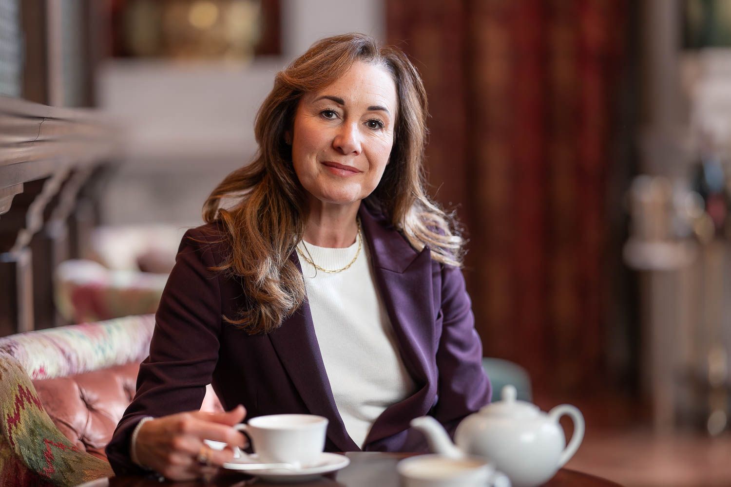 Environmental corporate portrait of a businesswoman in a plum jacket, seated with afternoon tea