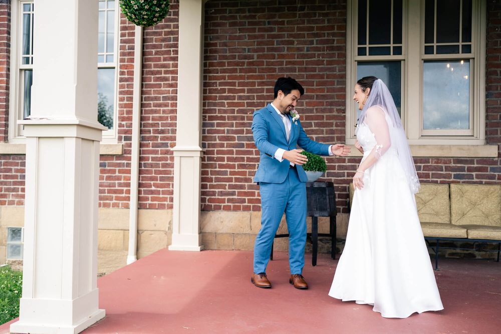 Groom in a blue suit holds a bouquet while a bride in a white gown and veil stands before him on a red porch of a brick house.