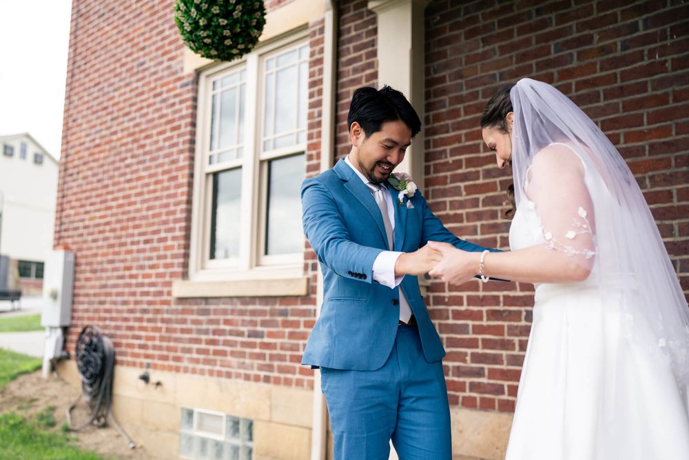 Groom in a blue suit holds hands with a bride in a white dress and veil beside a red brick building at their wedding.