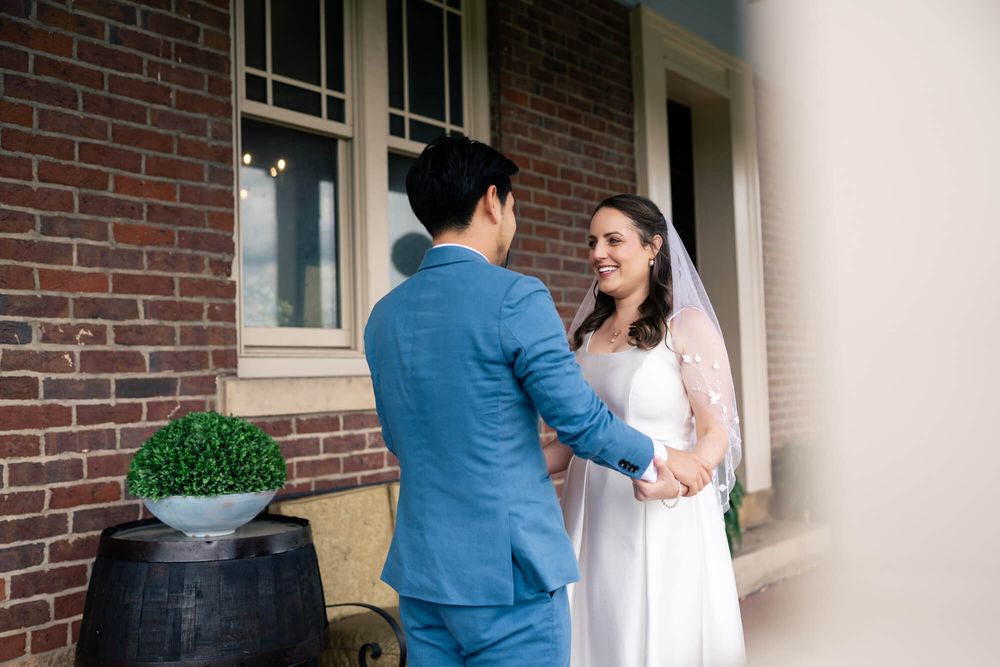 Smiling bride in a white dress and veil holds hands with a groom in a blue suit outside a brick building on their wedding day.