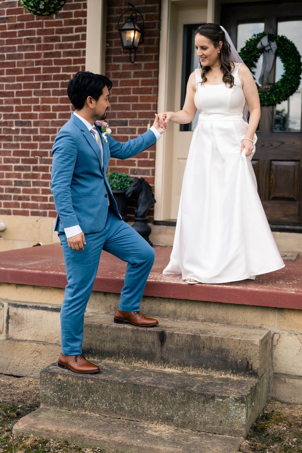 Groom in a blue suit and boutonniere steps up a stone staircase holding the hand of a smiling bride in a white wedding gown.