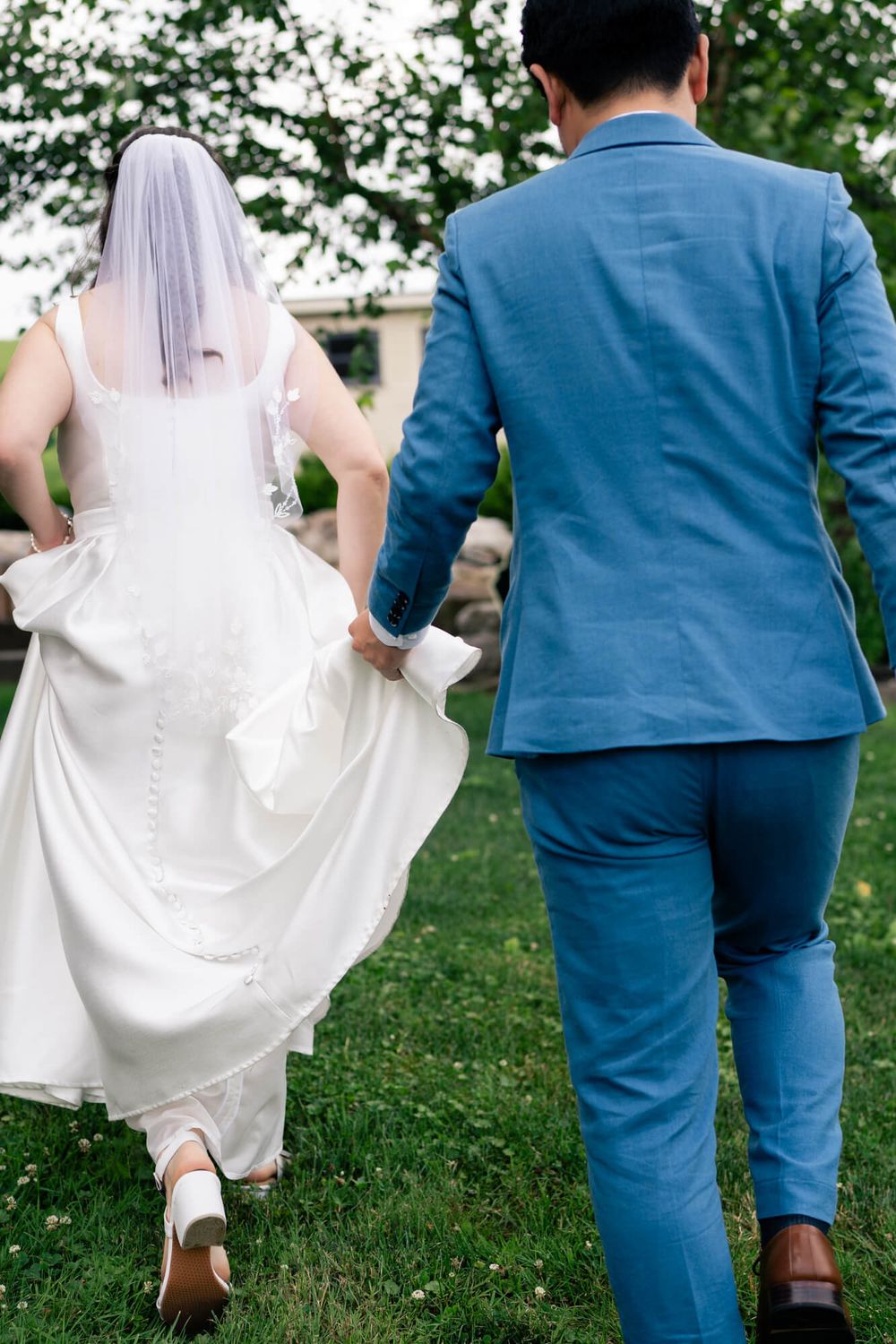 Bride in a white gown and veil lifts her skirt and walks across a green lawn holding hands with her groom in a blue suit.