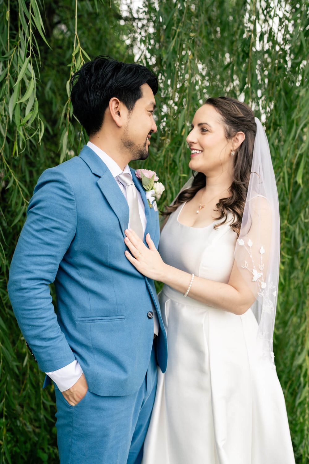 Smiling bride in a white gown and veil gazes at her groom in a blue suit in front of weeping willow branches at their wedding.