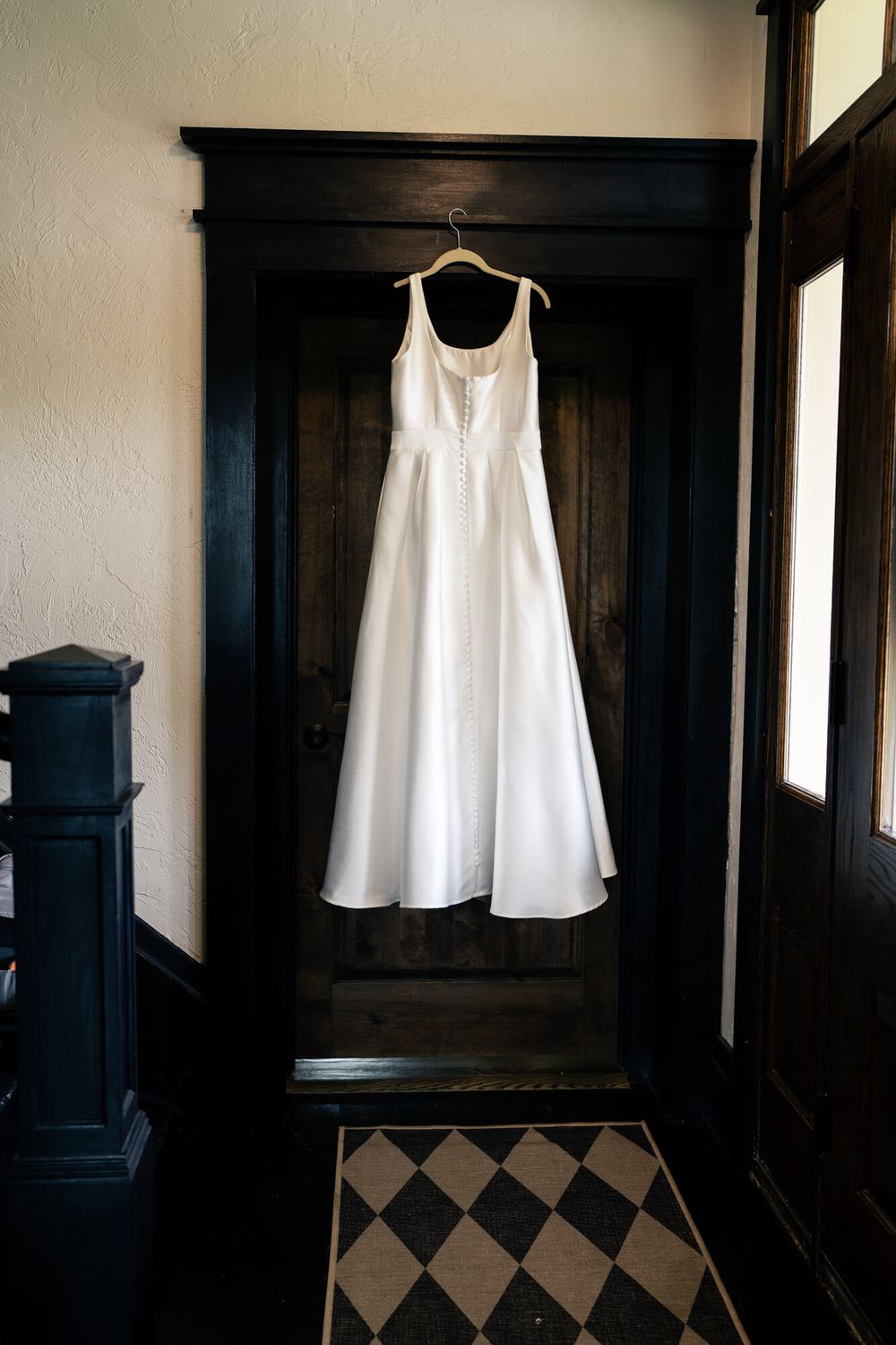 White sleeveless wedding dress with a button back hanging from a hook on a dark wooden door above a checkered tile floor.