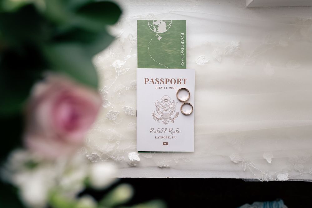 Two wedding rings rest on a passport-style invitation with a green cover, beside soft pink flowers and greenery on a lace tablecloth.