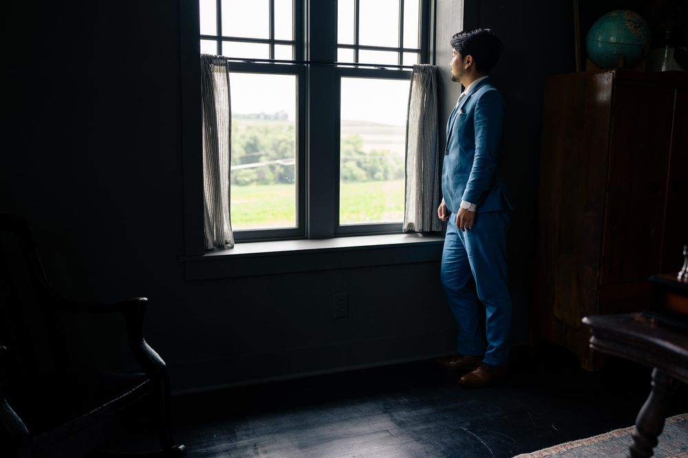 Person in a blue suit stands beside a window in a dim, dark room, gazing out at a sunlit green field and trees.