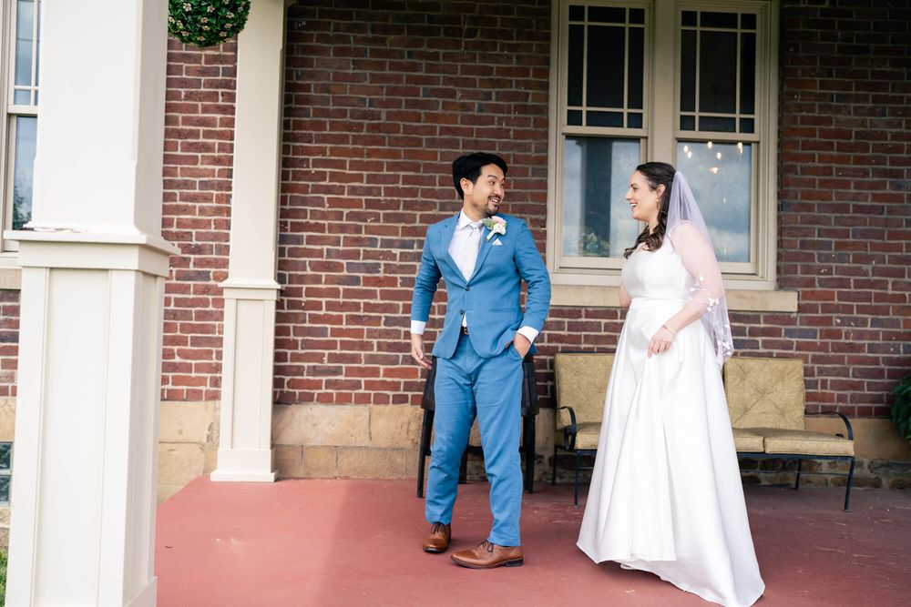 Groom in a blue suit and bride in a white gown with veil share a joyful moment on a red brick porch.
