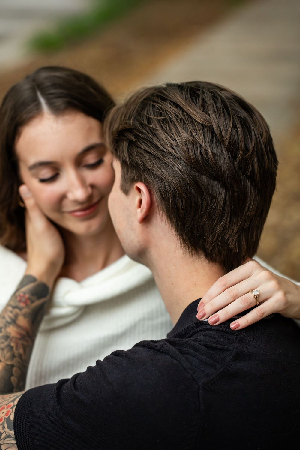 Smiling woman with a tattooed arm and a diamond ring gazes lovingly at a man in a black shirt as they embrace outdoors.