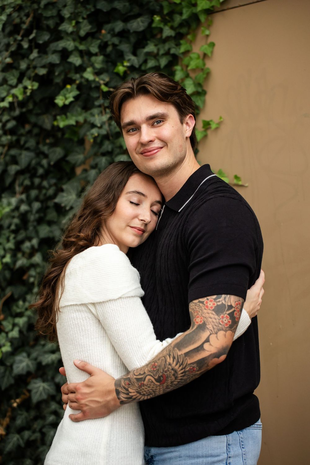 A smiling man with a tattooed sleeve embraces a woman with long brown hair resting her head on his chest beside an ivy-covered wall.