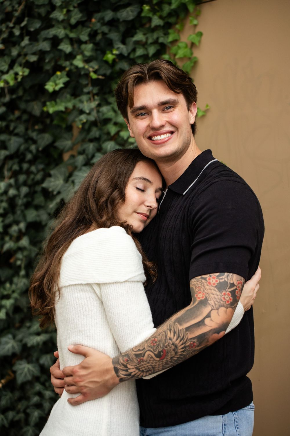 Smiling man with a forearm tattoo embraces a woman in a white sweater who rests her head on his chest in front of an ivy-covered wall.