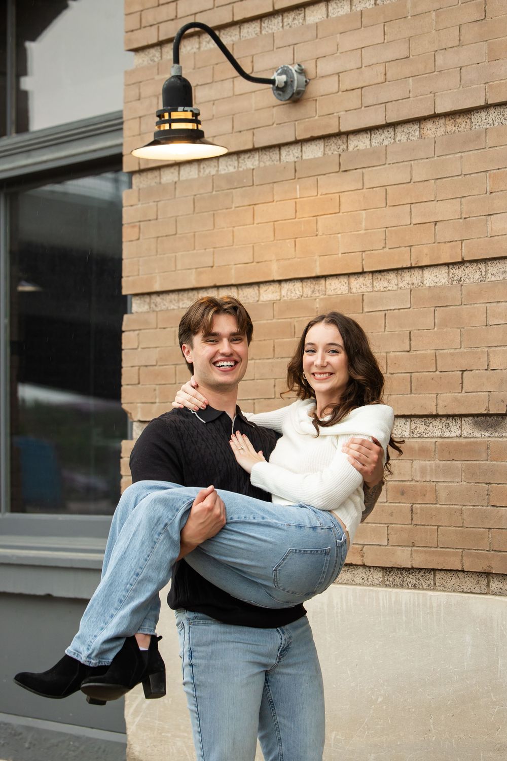 A smiling young man in a dark sweater carries a laughing woman in a white top and jeans in front of a tan brick wall with a barn light.