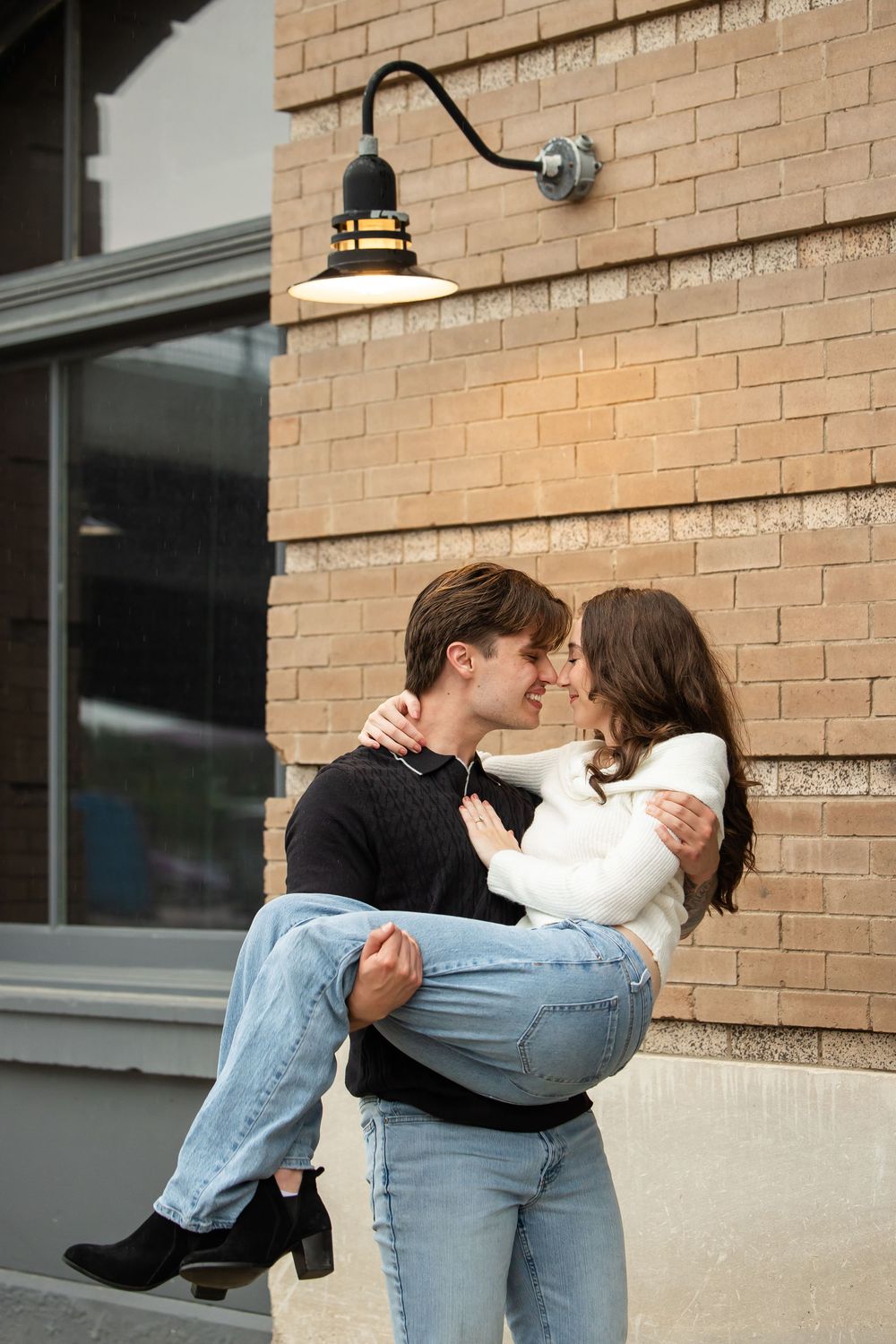 A man in a black sweater lifts a smiling woman in jeans and a white top, their noses touching, in front of a brick wall with a barn light.