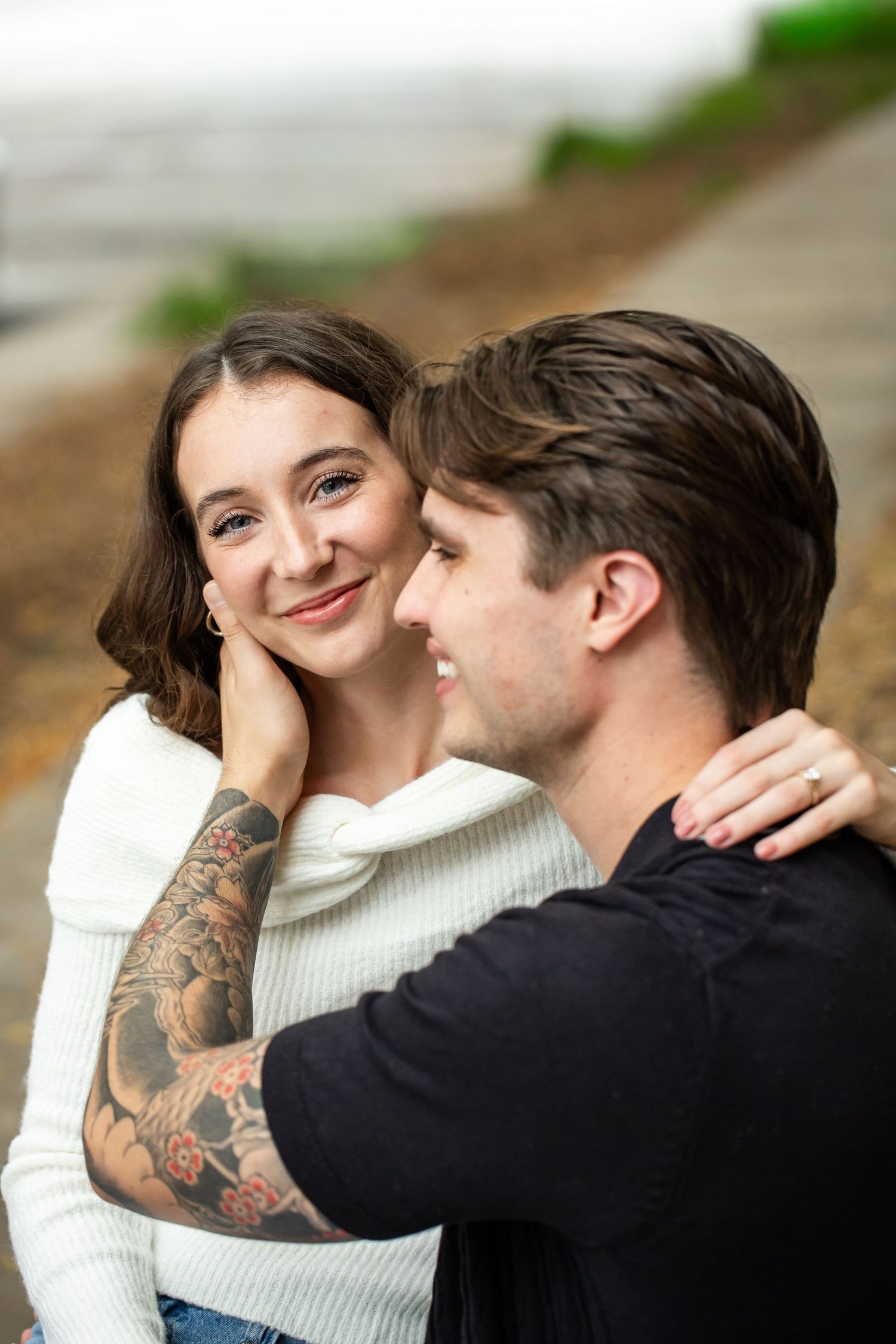 Smiling young woman in a white sweater rests her cheek against a man with a floral forearm tattoo in a warm outdoor embrace.