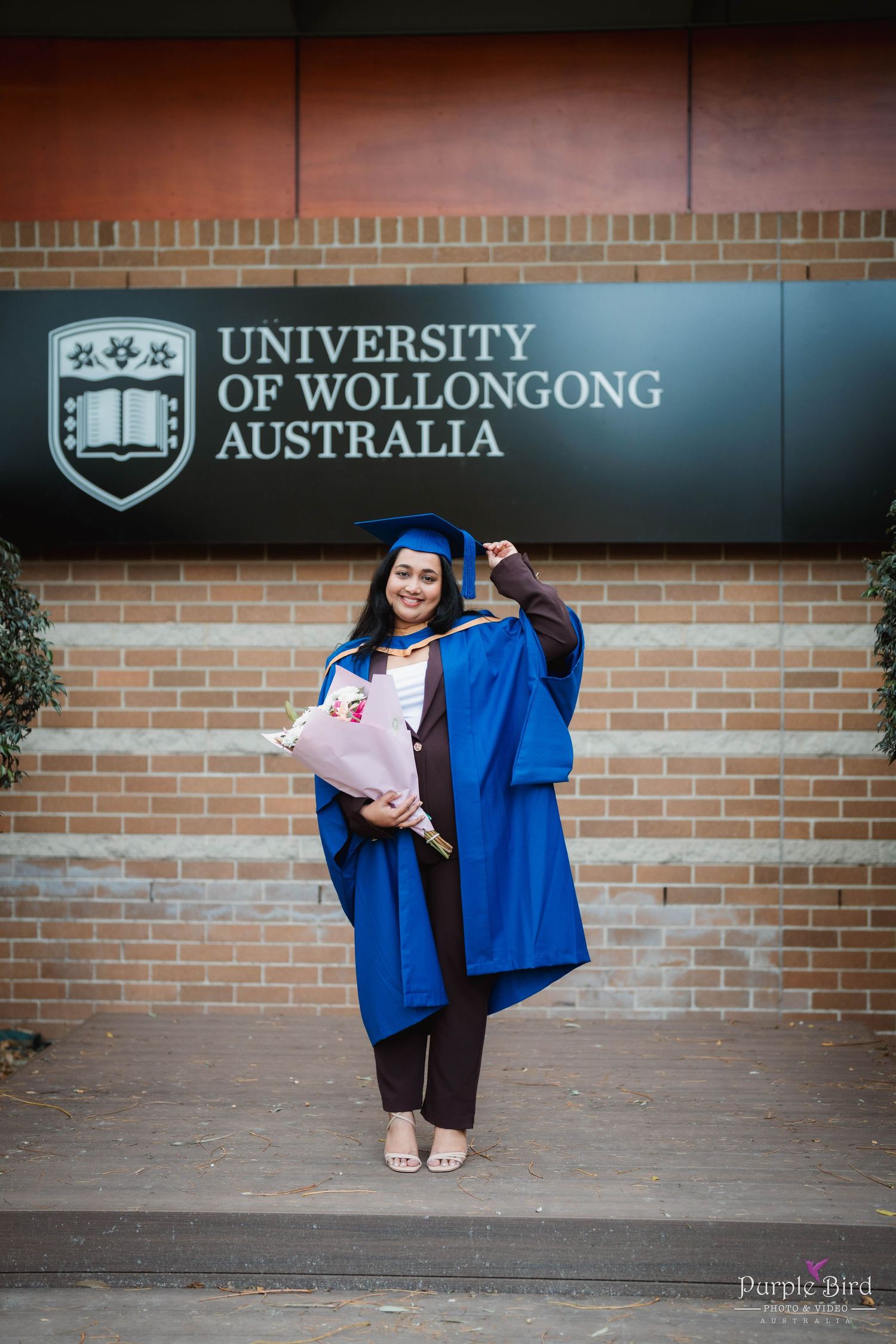 Graduate in blue gown and cap holding a bouquet while posing in front of the University of Wollongong Australia sign.