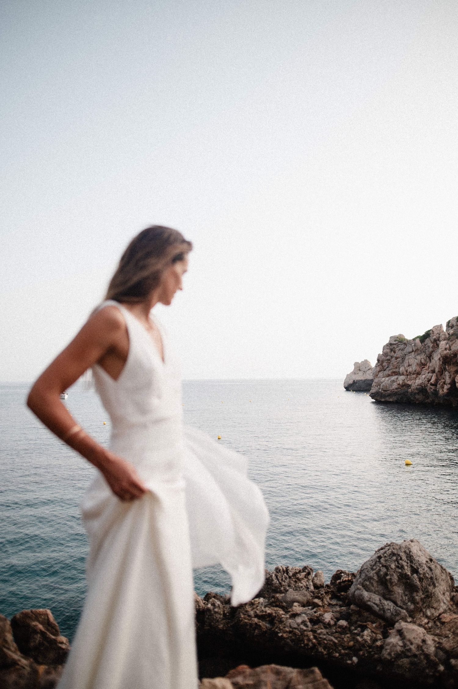 Woman in a flowing white dress stands on rugged coastal rocks overlooking calm blue sea, with soft focus and a hazy sky.