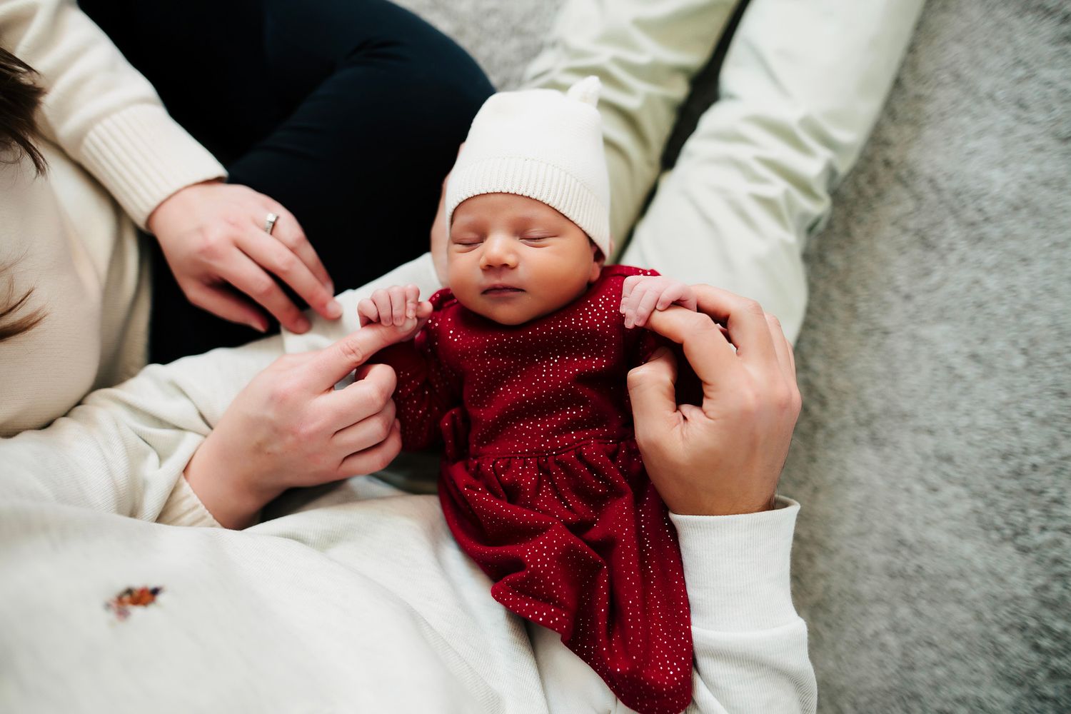 Parents tenderly dressing their sleeping newborn baby in a red knit outfit and white hat.