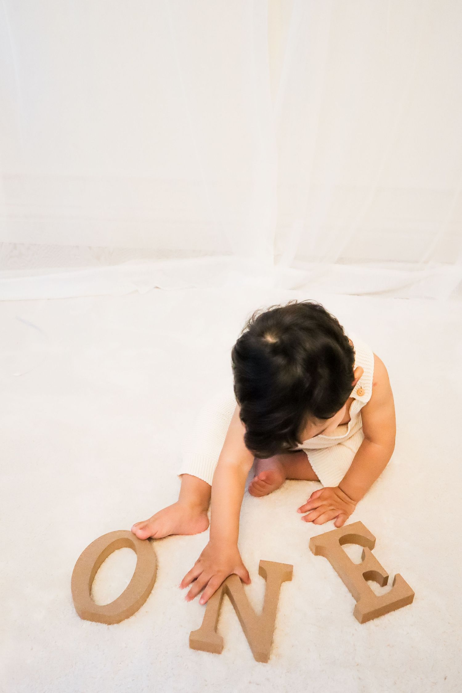 Baby reaching for wooden ONE letters on white floor, celebrating first birthday milestone.