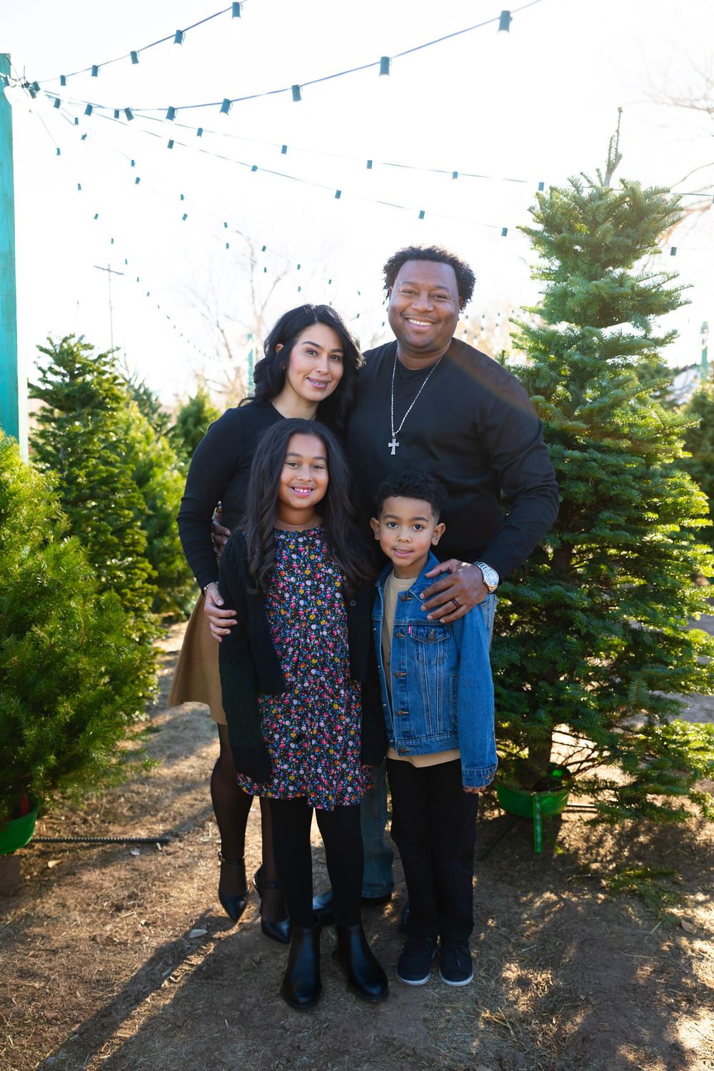 Family of four smiling together at a Christmas tree farm with string lights in the background.