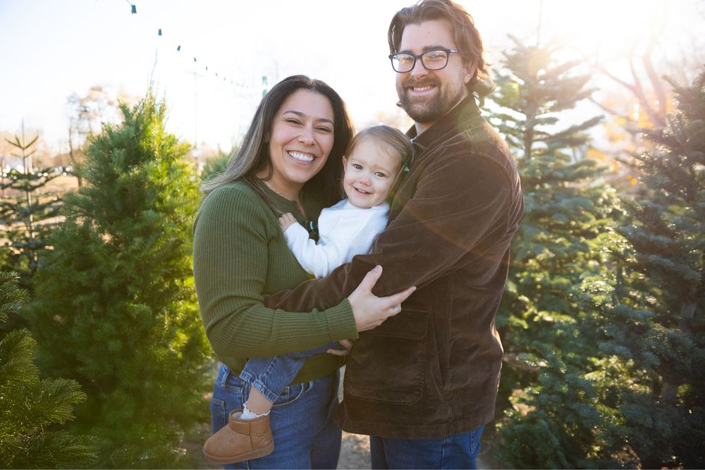 Happy family with baby posing at Christmas tree farm with evergreen trees in background on sunny day.