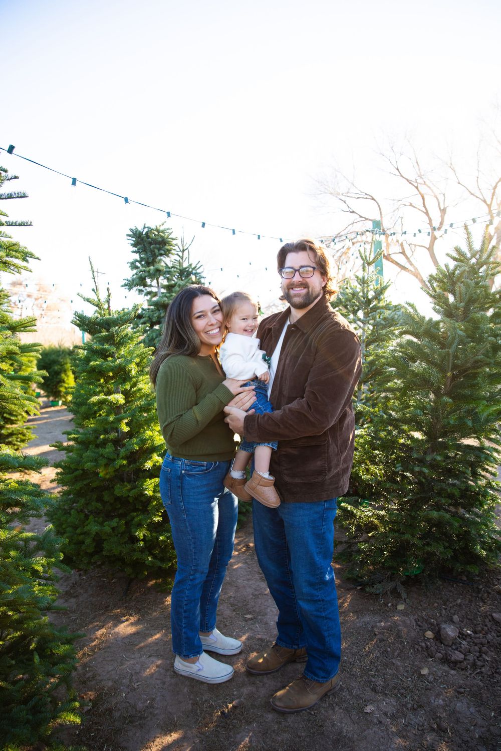 A happy young family with a baby poses among green Christmas trees at an outdoor tree farm with string lights.