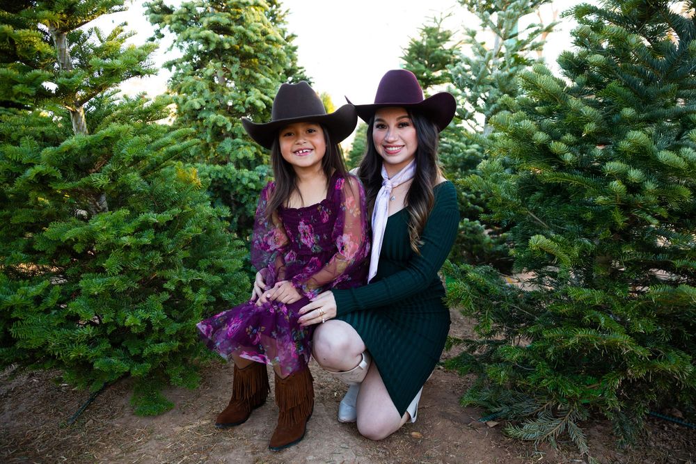 Two girls in cowboy hats and western outfits pose among green Christmas trees at a tree farm.